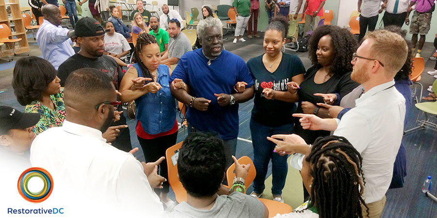 A group of educators, family members, and community partners participate in a group activity in a school library. RestorativeDC logo.